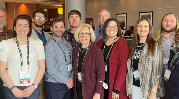 A group of 14 people pose for a photo at a conference.