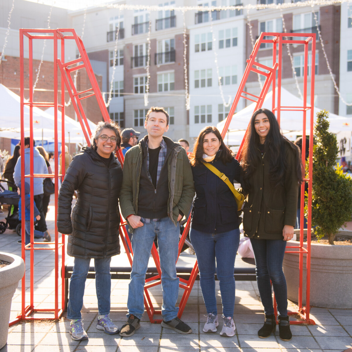 Four people stand shoulder to shoulder in front of a large red "M" structure. Behind them, a plaza is filled with visitors and white pop-up canopy tents staffed by vendors participating in a spring bazaar event.