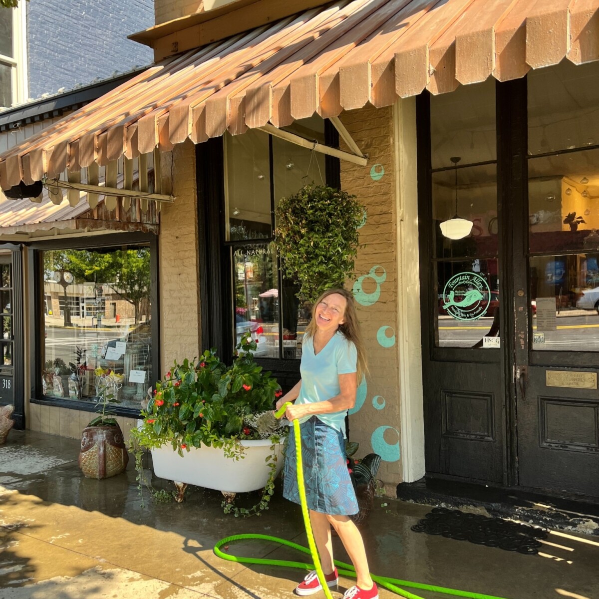 A woman smiles while watering plants in a vintage bathtub-turned-planter placed outside her store.