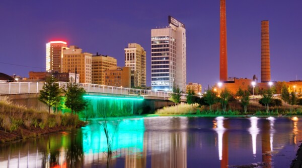 The underside of a bridge is illuminated with teal lights; the city skyline and rust-colored smoke stacks that rise in the background are reflected in the river that flows gently in the foreground.