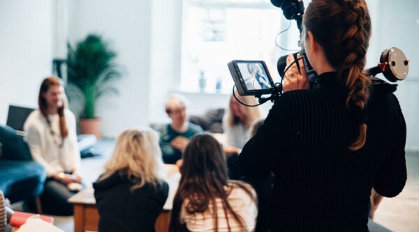 A woman holding a large camera films a group of people sitting around a table