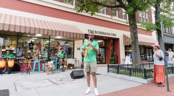 A saxophonist, drummer, and vocalist set-up on a wide sidewalk perform for pedestrians.