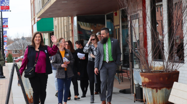 A small group of people walk down a sidewalk; one person smiles and waves while others engage in conversation.