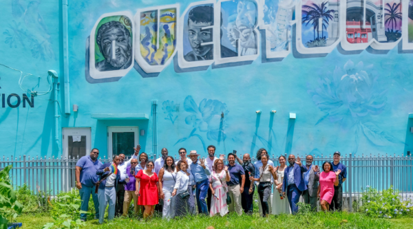 A group of people stand in front of exterior building mural that reads, "Overtown."