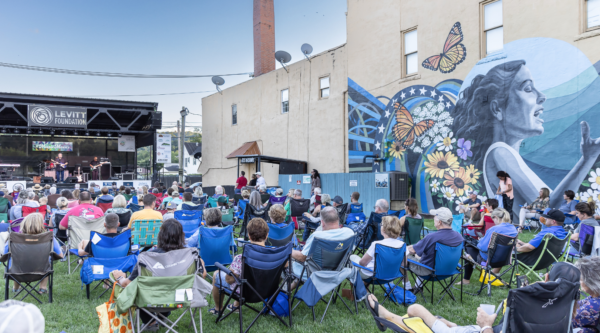 Group of people sit on lawn chairs on grassy lawn while listening to live music on outdoor stage next to mural on exterior of historic building.