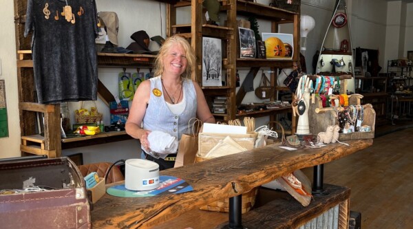 A woman smiles while standing behind a retail store counter and placing an item into a paper bag.