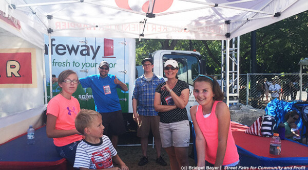 Three adults and three children smiling at the camera at an outdoor street fair.