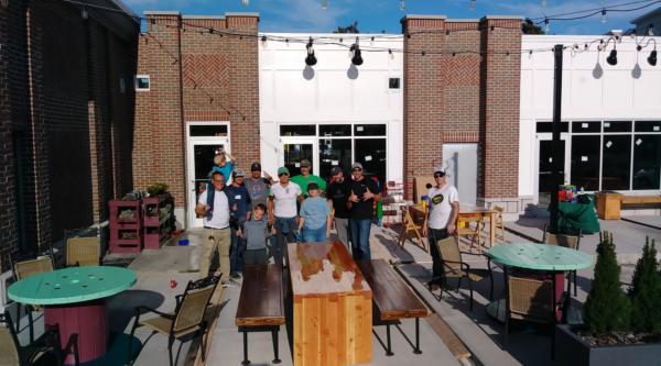 A group of people smile in public gathering space with tables, chairs, and strung lights.