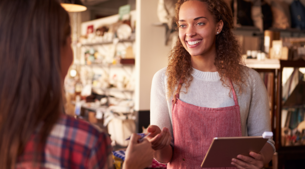 A woman in an apron holds an iPad and takes a credit card from a customer.