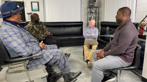 A group of four men sit in a circle talking in a barbershop.