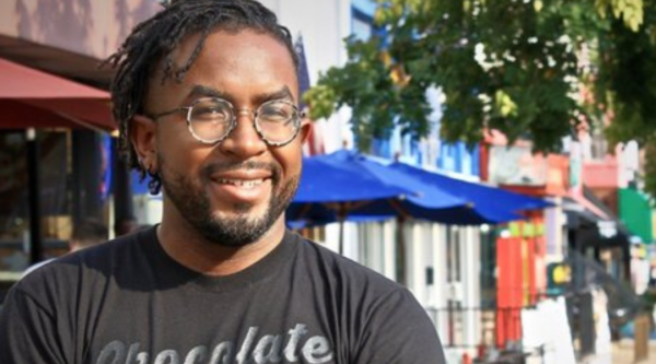 A man wearing glasses and a black shirt stands outside of small businesses with awnings.