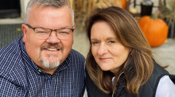 A man and woman, Glen Ellis and his wife Nancy Ellis, smile in front of doorway with autumn decorations.