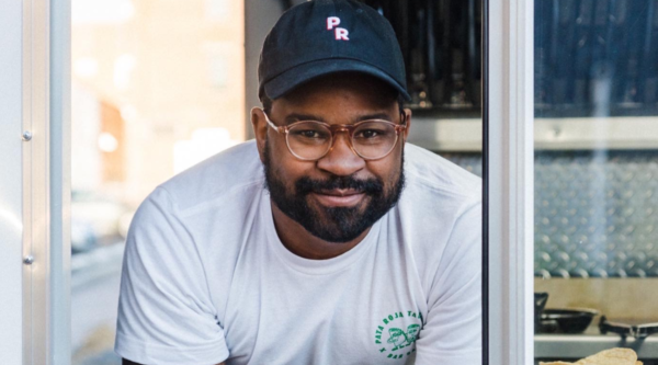 Photo of man wearing blue baseball cap and white t-shirt leaning out the window of a food truck.