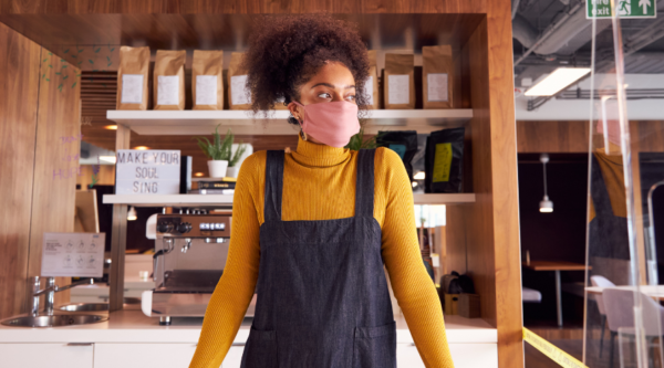 A woman wearing a medical face covering and an apron stands behind the counter in a coffee shop