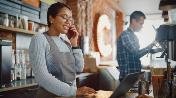 A woman standing at a cash register in a coffee shop holds a phone to her ear